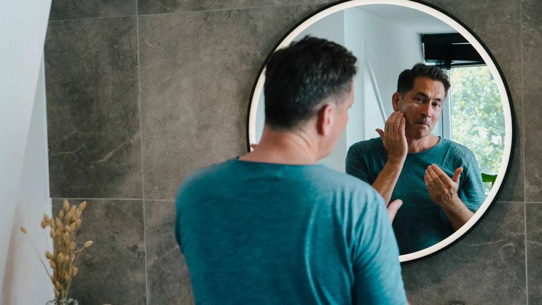 Man reinigt zijn gezicht voor een ronde badkamerspiegel en masseert de cleanser in op zijn wang