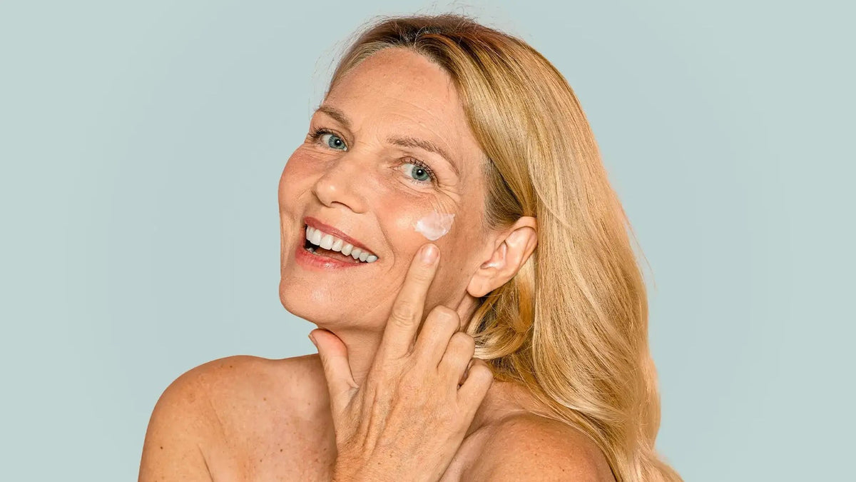 Vrouw brengt lachend dagcrème met SPF aan, haar voorhoofd vrij in het licht